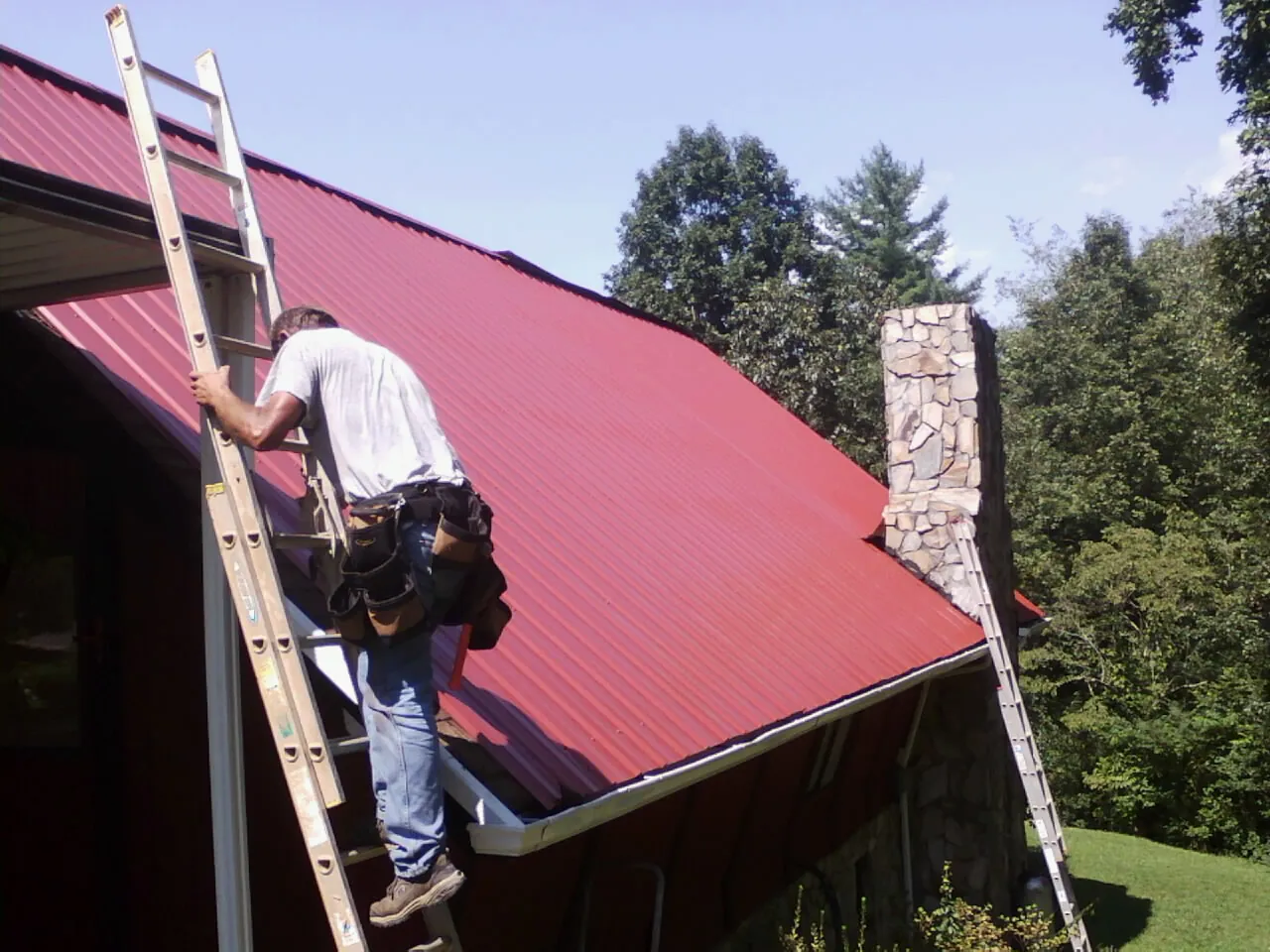 Roofer on a ladder beside a red metal roof and stone chimney — Soffit & Fascia Repair by Roofing Brothers in Pacoima